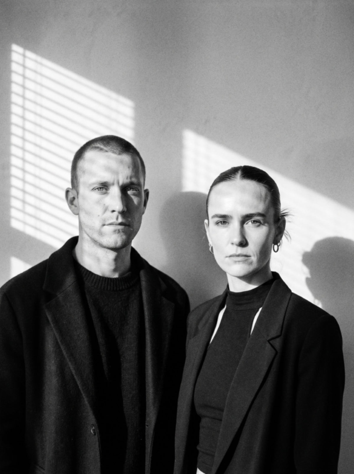A black and white portrait of a man and a woman standing side by side against a wall with strong window shadow patterns. Both are dressed in dark, minimalist outerwear and knit tops.